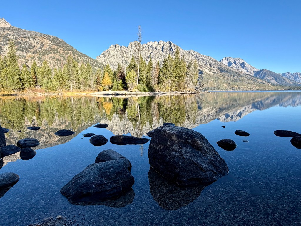 Calm lake reflecting mountains and trees under a clear blue sky, with rocks in the foreground.