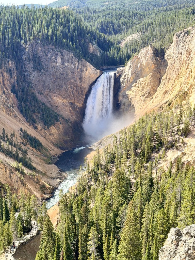 A stunning view of a large waterfall cascading down steep cliffs, surrounded by dense green forests and rocky terrain in a mountainous landscape.